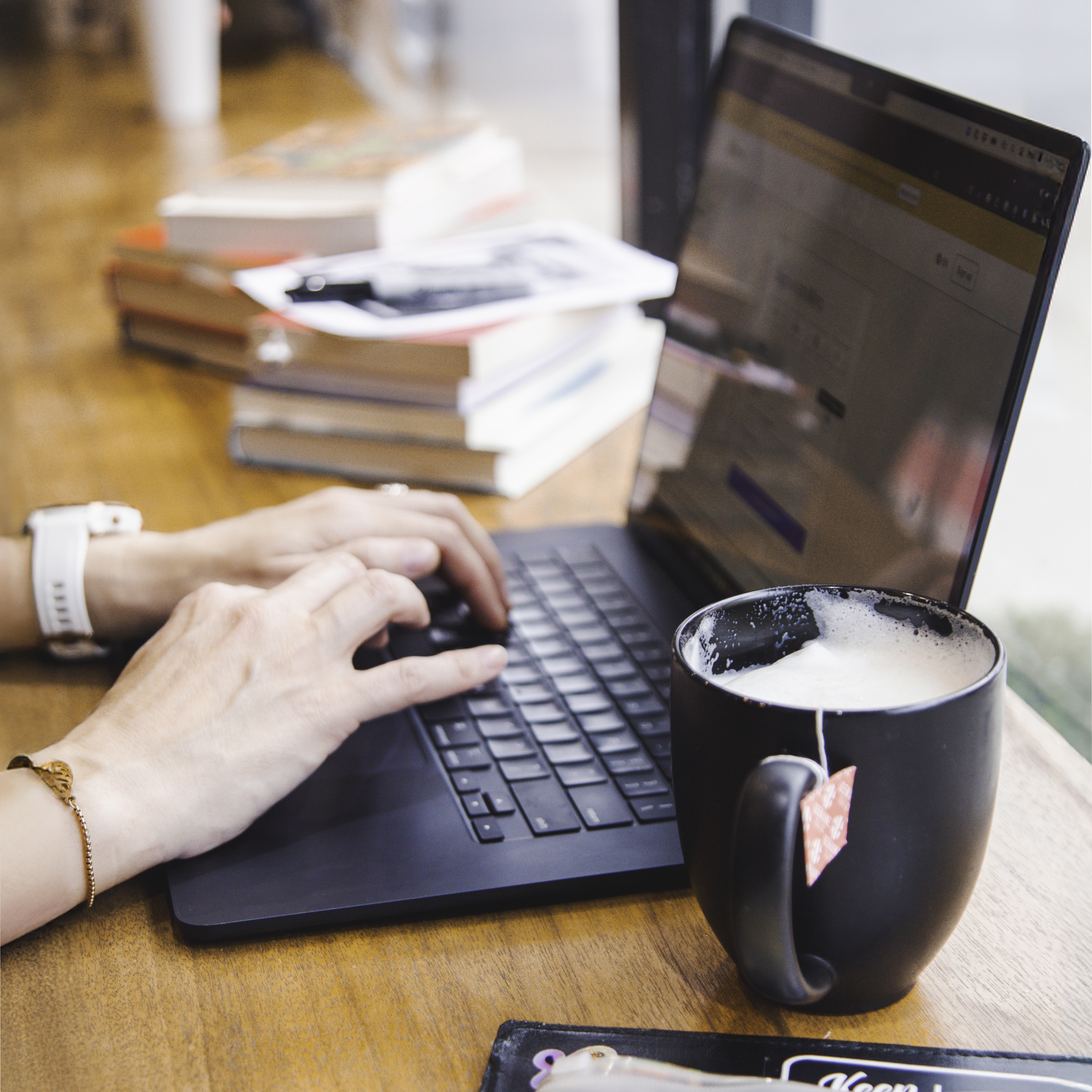 Laptop with hands on keyboard, a cup of tea, and stacks of books nearby